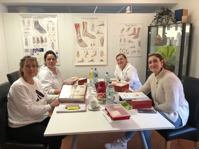 Vier Teilnehmerinnen am Schulungstisch mit Lehrmaterialien und anatomischen Lehrtafeln im Hintergrund